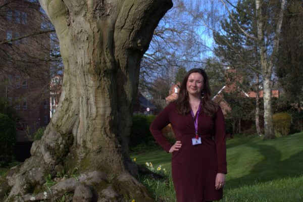 Hollie stands to the right of a large tree in the Hospice grounds. She's wearing a burgundy dress and gold earrings. Spring flowers are in the background.