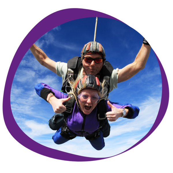 two people taking part in a tandem skydive. The background is a bright blue sky with light wispy clouds.