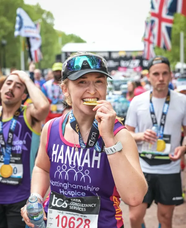 Marathon participant, wearing a purple Nottinghamshire Hospice running top with the name 'Hannah' on the front, bites a gold-colored medal at the finish line of a marathon, with other runners in the background.