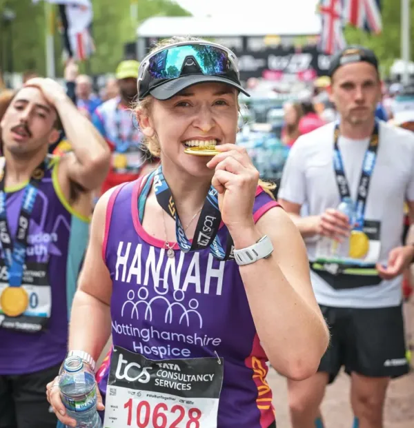 Marathon participant, wearing a purple Nottinghamshire Hospice running top with the name 'Hannah' on the front, bites a gold-colored medal at the finish line of a marathon, with other runners in the background.