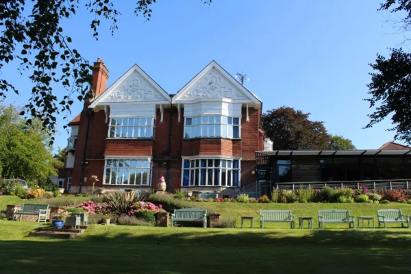 Fernleigh House, a large red-brick building with white decorative gables and wide bay windows, home to Nottinghamshire Hospice and Portland College. The building is surrounded by landscaped gardens with colorful flowers, green lawns, and garden benches under a clear blue sky.