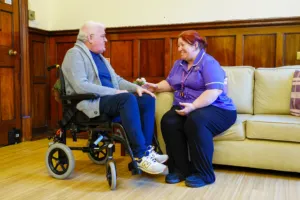 Hospice care worker in a purple uniform smiling  with a man in a wheelchair indoors.