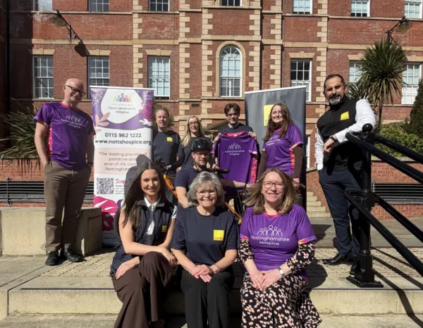 A group of people pose outdoors in front of a red‑brick building with large windows and tall plants. Several wear purple “Nottinghamshire Hospice” shirts, while others wear dark shirts with yellow badges. Two banners stand behind them: one for Nottinghamshire Hospice with contact details and a tagline about palliative and end‑of‑life care, and another featuring the Savills logo. The group appears to be gathered for a community or charity event.