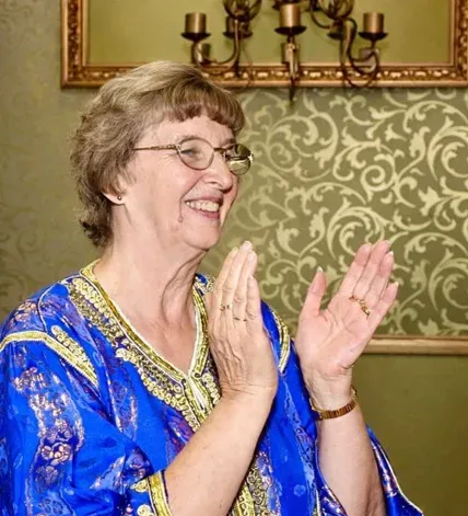 A smiling woman in a bright blue and gold blouse clapping her hands.